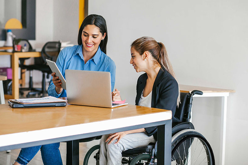 Young woman in a wheelchair sitting with her female colleague, next to a computer at workplace.
